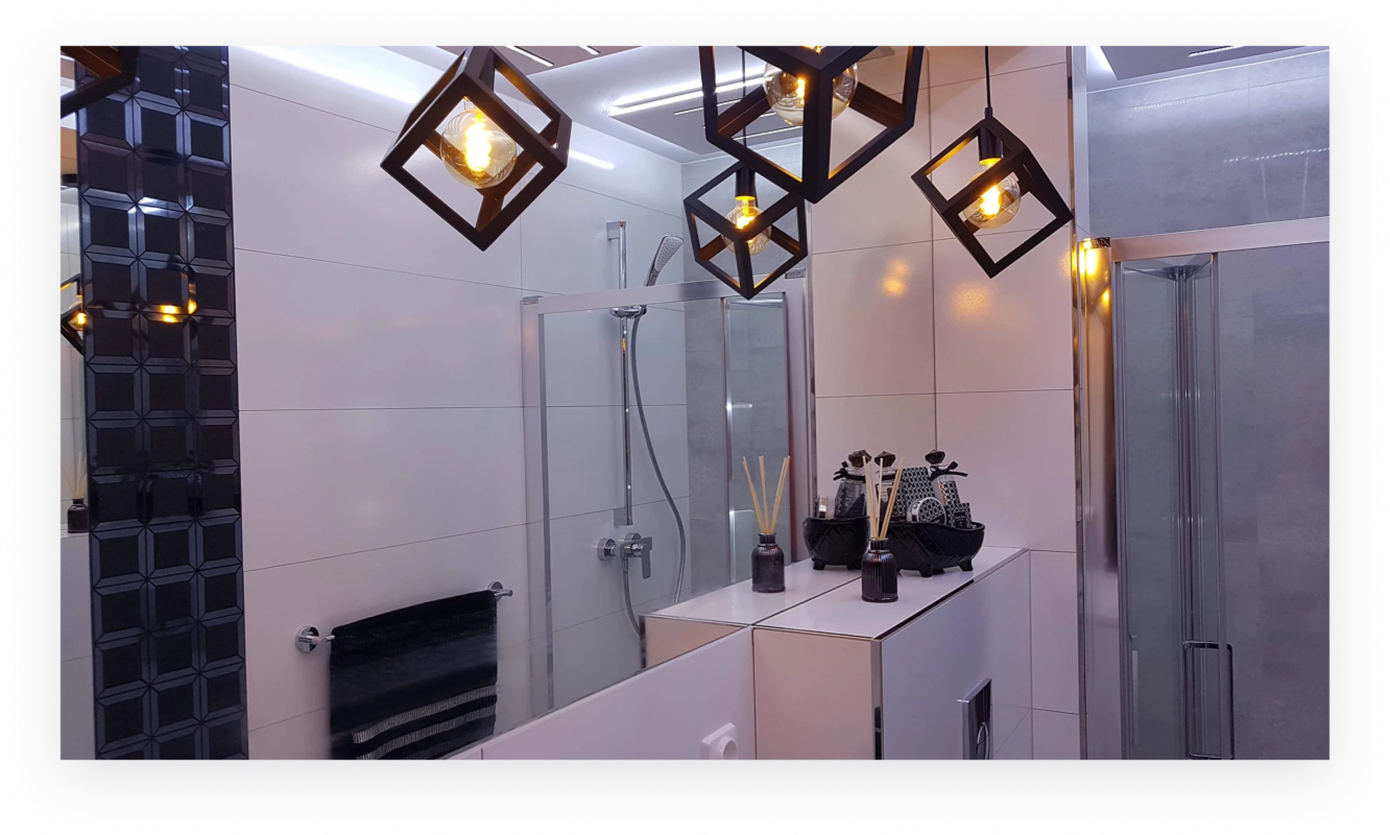 Aranżacja łazienki z białymi płytkami, czarnymi akcentami, szklaną kabiną prysznicową i geometrycznymi lampami