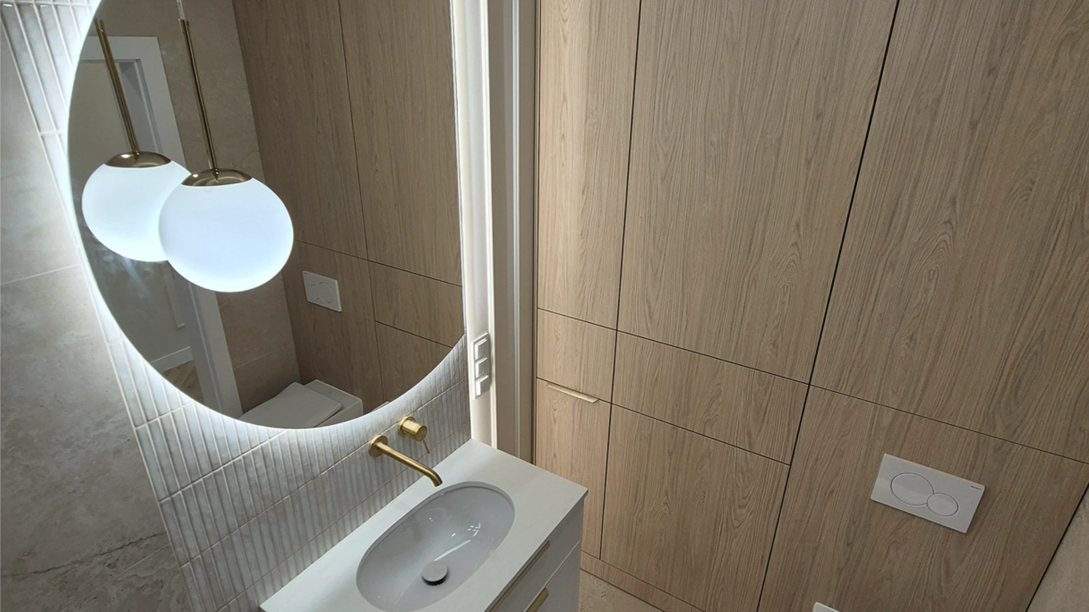 Aranżacja łazienki: umywalka z złotym kranem, podświetlane okrągłe lustro i drewniana zabudowa ścian