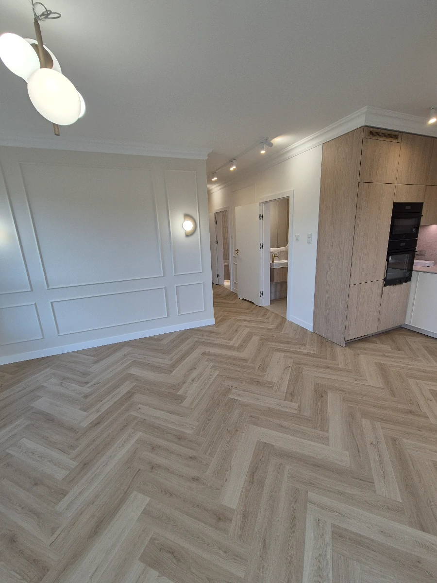 Jasne wnętrze z podłogą ułożoną w jodełkę (układanie paneli podłogowych), zabudową kuchenną i sztukaterią ścienną