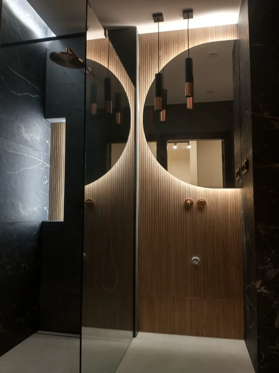 Aranżacja łazienki z podświetlanym okrągłym lustrem, drewnianymi panelami i czarnym marmurowym wykończeniem